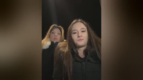 Media: Video of two young women with long brown hair, wearing black jackets, walking side by side in a dimly lit corridor, with blurred, out-of-focus background.