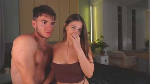 Media: Video of a shirtless, muscular man and a brunette woman in a modern, dimly lit living room. The woman covers her mouth with her hand, and both appear to be in a private moment.