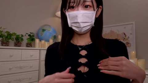 Media: Video of an East Asian woman in a black sweater with cutouts, wearing a white mask and gloves, giving a thumbs-up gesture, in a home setting with a globe, potted plants, and candles on a dresser.