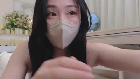 Media: Video of an Asian woman with long black hair, wearing a beige mask, and a white top, taking a selfie indoors. Background features a white shelf with green plants and a globe.