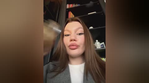 Media: Video of a fair-skinned woman with long brown hair, wearing a grey cardigan over a white shirt, puckering her lips for a kiss. Background includes a dark bookshelf with books and decorative items.