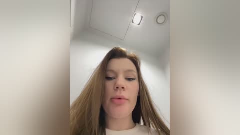 Media: A video of a young woman with light skin, long brown hair, and a white top, making a pouty face. She stands in a white-walled, ceiling-lit room with a rectangular light fixture.