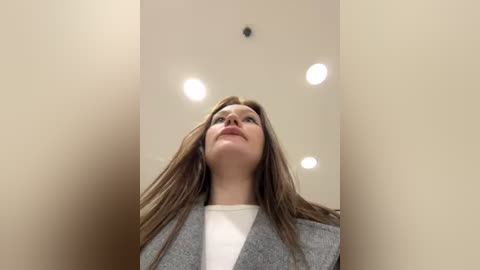 Media: Video of a young woman with long brown hair, wearing a gray blazer over a white shirt, looking upwards in a beige-walled room with recessed ceiling lights.