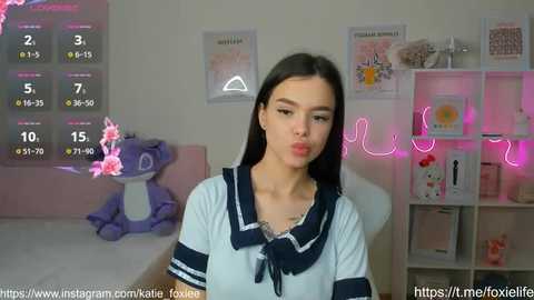 Media: Video of a young woman with fair skin, long dark hair, and a white sailor collar shirt, seated in a pastel-themed room with pink neon lights and plush toys.