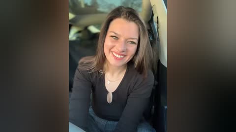 Media: Video of a smiling, light-skinned woman with shoulder-length brown hair, wearing a black V-neck top and jeans, seated in the passenger seat of a car.