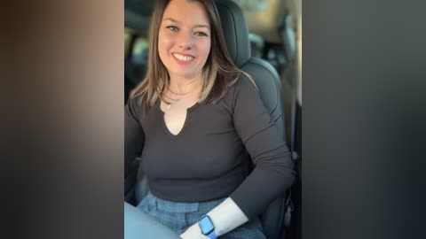 Media: A video of a young woman with straight, shoulder-length brown hair, wearing a black V-neck top and blue jeans, sitting in a car seat, smiling.