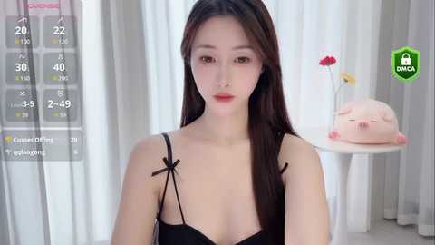 Media: A video of an East Asian woman with fair skin and long black hair, wearing a black spaghetti strap top, standing in a bright, minimalist room with white curtains and a pink pig figurine.