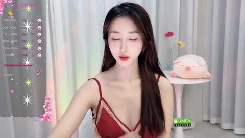 Media: Video of a young Asian woman with long black hair in a red lace bra, smiling, in a softly lit room with white curtains and a pink pillow.