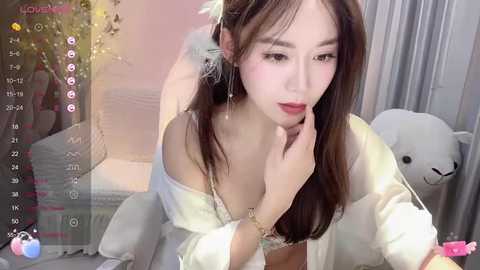 Media: Video of an Asian woman with long brown hair, wearing off-shoulder lingerie, touching her face, in a soft, pastel bedroom with a teddy bear and a plush pillow.