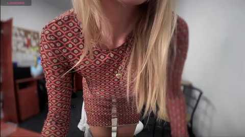 Media: Video of a slender, blonde woman with long hair, wearing a red patterned blouse and white shorts, leaning forward in an office setting.