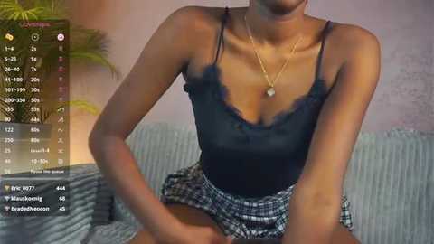 Media: Video of a slender, light-skinned woman with a small bust, wearing a black lace camisole and plaid skirt, sitting on a sofa. Background shows a potted plant and a soft, pastel wall.