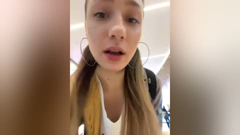 Media: A candid video of a young woman with long blonde hair, light skin, and hoop earrings, wearing a white top and mustard-yellow jacket, taken from a close-up, slightly blurry perspective.