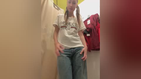 Media: Video of a young woman with light skin and long blonde pigtails, wearing a white t-shirt with a graphic and ripped blue jeans, standing in a fitting room with red and beige clothing hanging nearby.