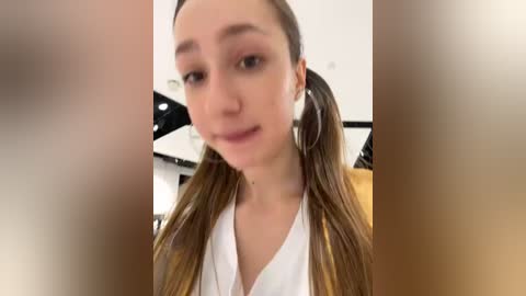 Media: A blurry video of a young woman with light skin, long brown hair in pigtails, wearing a white top, and large hoop earrings, taken indoors with a modern background.