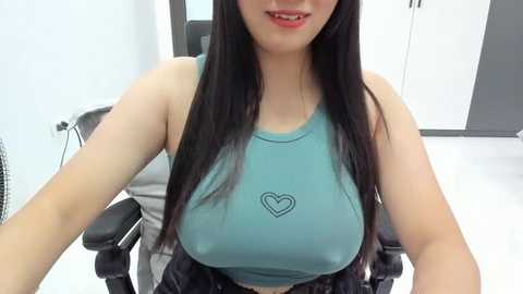 Media: Video of an East Asian woman with long black hair, fair skin, wearing a turquoise sports bra and patterned skirt, sitting on a black office chair in a modern, white-walled office.