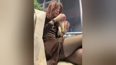 A video of a woman with shoulder-length brown hair, wearing a black dress and black stockings, sipping a coffee while seated on a white couch. The background includes a glass window and potted plant, suggesting an indoor setting.