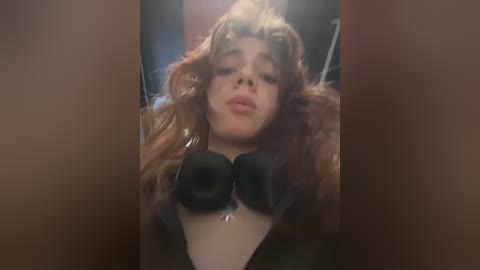 Media: Video of a young woman with wavy, light brown hair and fair skin, wearing a fur-trimmed coat, black headphones, and a silver necklace, blowing a kiss.