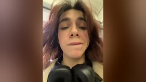 Media: Video of a young woman with messy, shoulder-length brown hair, wearing a black top and large black headphones, looking slightly annoyed.