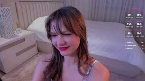 Media: Video of a smiling Asian woman with long brown hair, wearing a white tank top, in a modern, softly lit bedroom.