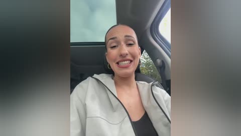 Media: Video of a smiling, light-skinned woman with a shaved head, wearing a white and black jacket, sitting in a car with a cloudy sky visible through the window.