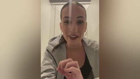 Media: Video of a smiling, light-skinned woman with medium-length braided hair, wearing a light gray jacket and black top, posing in a narrow, beige hallway with a glass wall.