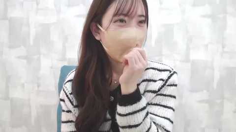 Media: Video of an Asian woman with long brown hair, wearing a white and black striped sweater, a beige mask, and a black dress, holding her hand to her mouth. She sits against a white and gray patterned curtain background.