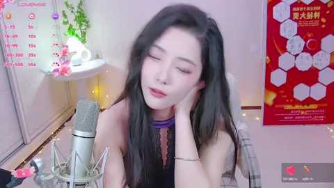 Media: Video of an Asian woman with long black hair, wearing a black lace top, leaning on a desk, with a microphone, calendar, and plants in the background.