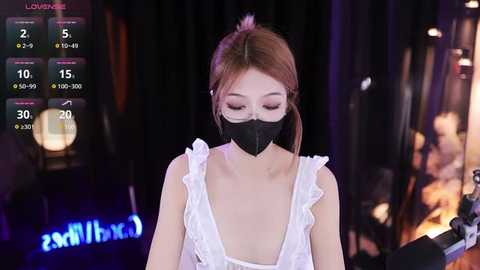 Media: Video of a young Asian woman with fair skin, brown hair, wearing a white sleeveless dress and black face mask, standing in a dimly lit room with a digital display showing temperature and humidity.