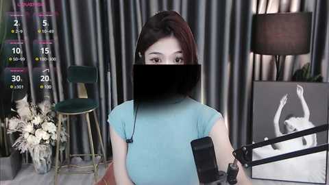 Media: Video of a young Asian woman with a medium build, wearing a blue t-shirt, seated at a desk with a microphone. Background includes gray curtains, a green chair, a flower arrangement, and a lamp.