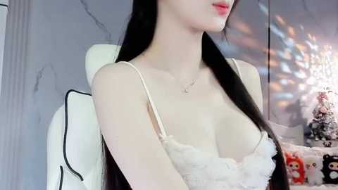 Media: Video of an Asian woman with fair skin and long black hair, wearing a white lace bra, sitting on a white gaming chair in a dimly lit room with a chandelier and colorful light reflections on the wall.