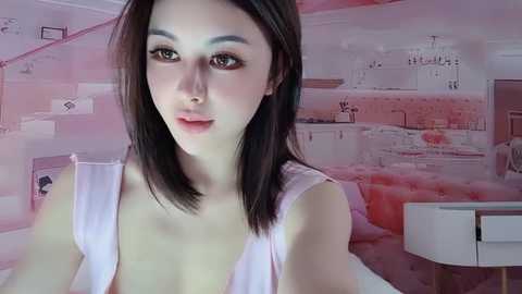 Media: A video of a young East Asian woman with straight, shoulder-length black hair and fair skin, wearing a sleeveless white top, in a modern, minimalist bedroom with pink and white decor.