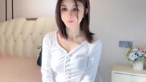 Media: Video of a young Asian woman with straight, shoulder-length dark hair, wearing a white button-up blouse, standing in a modern bedroom with a white tufted headboard, light-colored bedding, and a white nightstand with a floral arrangement.