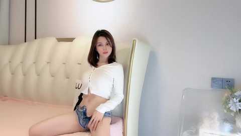 Media: Video of a slender Asian woman with straight brown hair, wearing a white cropped top and denim shorts, sitting on a cream bed in a modern bedroom.