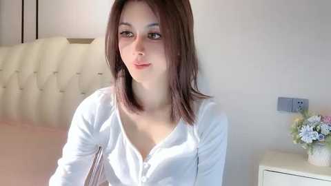 Media: Video of a young woman with shoulder-length brown hair, fair skin, and light makeup, wearing a white blouse, sitting on a beige couch with a tufted headrest in a softly lit, modern bedroom.