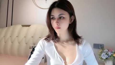 Media: Video of a young Asian woman with straight dark hair, wearing a white, low-cut, long-sleeved top, sitting in a cream-colored tufted headboard bedroom.