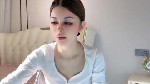 Media: Video of a young Asian woman with fair skin, dark hair in a ponytail, wearing a white blouse, sitting on a beige tufted headboard in a softly lit room.