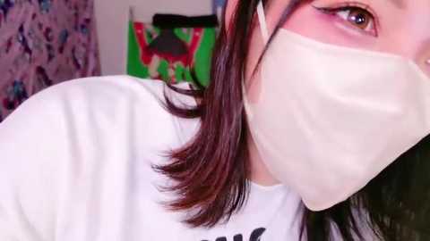 Media: A close-up video of a woman with fair skin and brown hair, wearing a white face mask and a white shirt, taken indoors with a blurred background of colorful, patterned fabric.