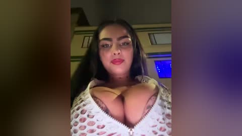 Media: Video of a dark-skinned woman with long, straight hair, wearing a white, lace-up top that accentuates her large breasts. She has a neutral expression and is indoors, with a dimly lit background.