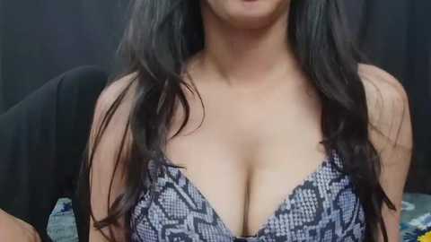 Media: Video of a woman with long, wavy black hair, wearing a revealing, snake-patterned black and white bra that accentuates her ample cleavage. Background features a dark, textured wall.