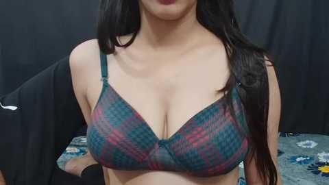 Media: Video of a woman with long dark hair, wearing a plaid-patterned, blue-green bra with underwire, black t-shirt draped over her shoulders, and a black jacket. Background is a dark curtain.