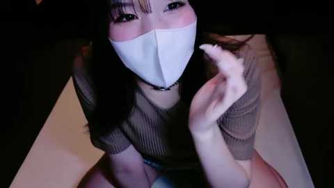 Media: Video of an Asian woman with long dark hair and pale skin, wearing a white surgical mask and a brown knit sweater, sitting on a white surface, finger pointing.