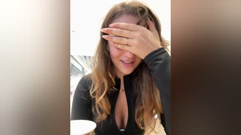 Media: Video of a smiling woman with long, wavy brown hair, wearing a black long-sleeve top, covering her eyes with her hand, sitting at a table with a white cup.