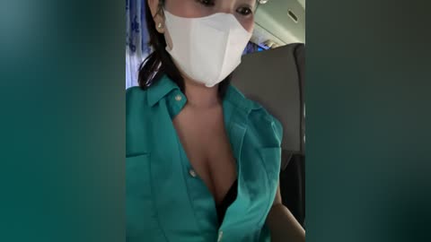 Media: Video of a woman in a teal nurse's uniform, with a white surgical mask covering her mouth and nose, seated in a car. Her dark hair is tied back, and her collarbone is partially visible.