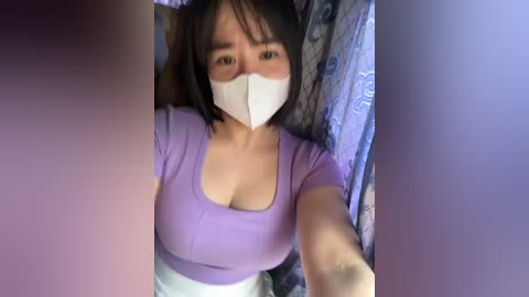 Media: A video shows an Asian woman with short black hair and light skin wearing a white mask, lavender top, and white pants, lying on a bed with blue and white patterned sheets.