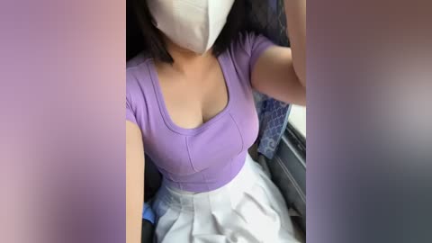 Media: Video of a woman in a purple top, white mask, and gray skirt, seated on a bus with a blurred background.