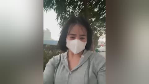 Media: Video of an Asian woman with dark hair and fair skin wearing a white face mask and gray hoodie, standing outdoors under a leafy tree, blurred background.