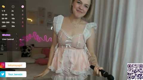 Media: Video of a young woman with blonde hair wearing a frilly, light pink lingerie set, holding a microphone. Background shows a cozy bedroom with a bed and a cat.