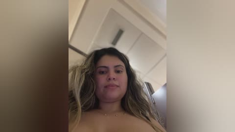 Media: A video of a young, light-skinned woman with long, wavy, blonde hair, partially covering her breasts. She has a neutral expression and is indoors, possibly in a bedroom.