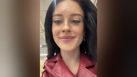 Media: Video of a young woman with straight black hair, wearing a red leather jacket, smiling. Background shows a blurred interior with beige and white walls.