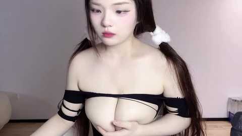 Media: A video of an East Asian woman with long black hair in pigtails, wearing a black off-shoulder top, revealing her large breasts, in a minimalist room with a beige wall and wooden floor.